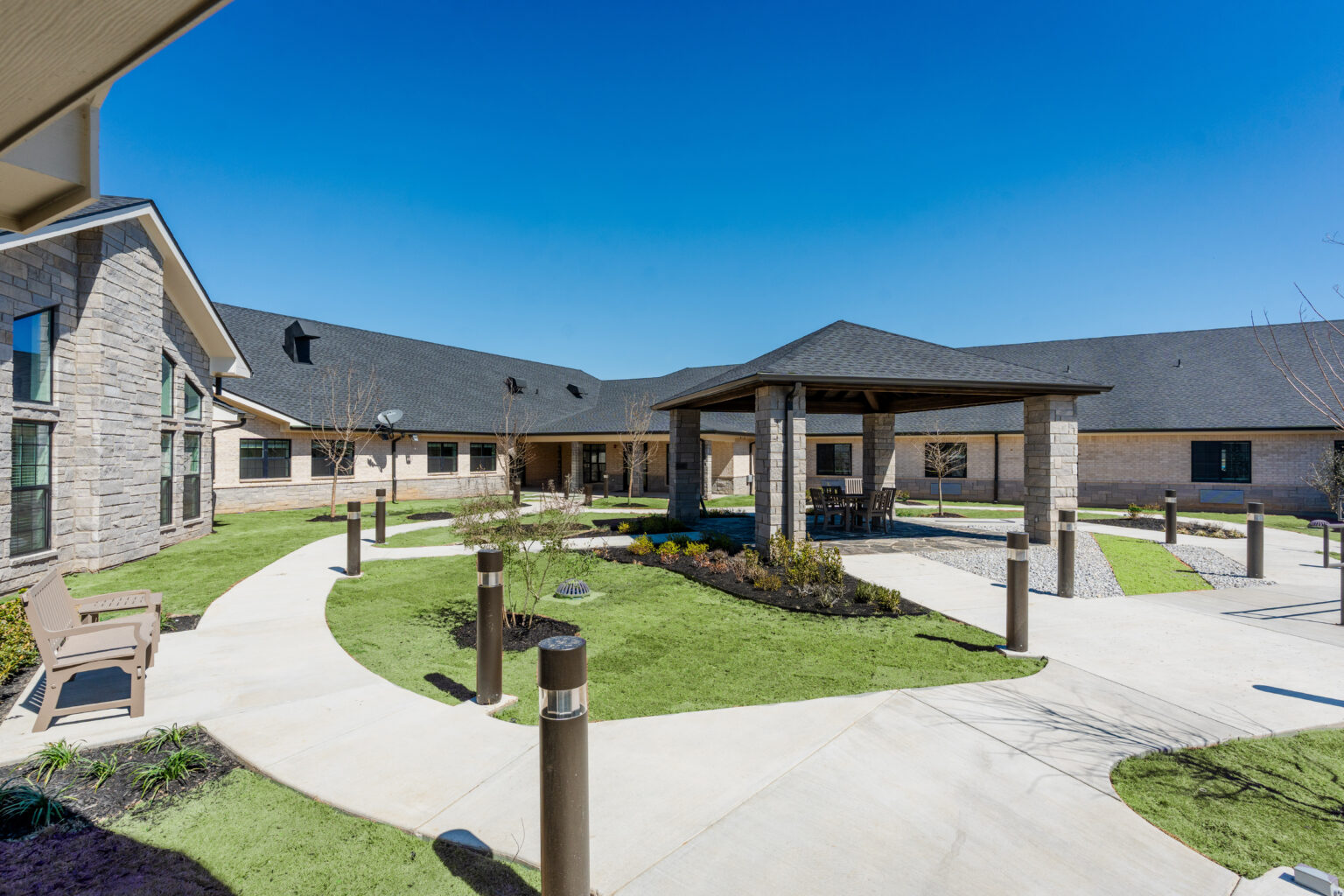 The exterior Courtyard at Quail Run Health and Rehab, a modern nursing home and elderly care center. The building features a covered pergola area, and a lighted walking trail. This skilled nursing and senior living facility offers an environment for residents seeking Memory care.