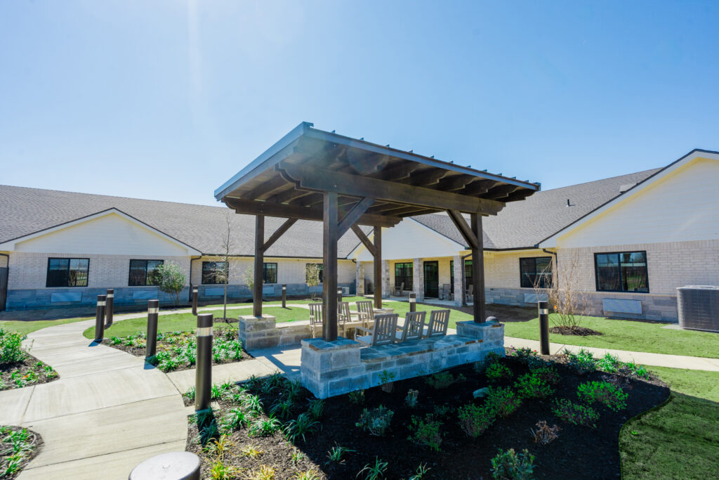 The exterior Courtyard at Quail Run Health and Rehab, a modern nursing home and elderly care center. The building features a covered pergola area, and a lighted walking trail. This skilled nursing and senior living facility offers an environment for residents seeking Memory care.